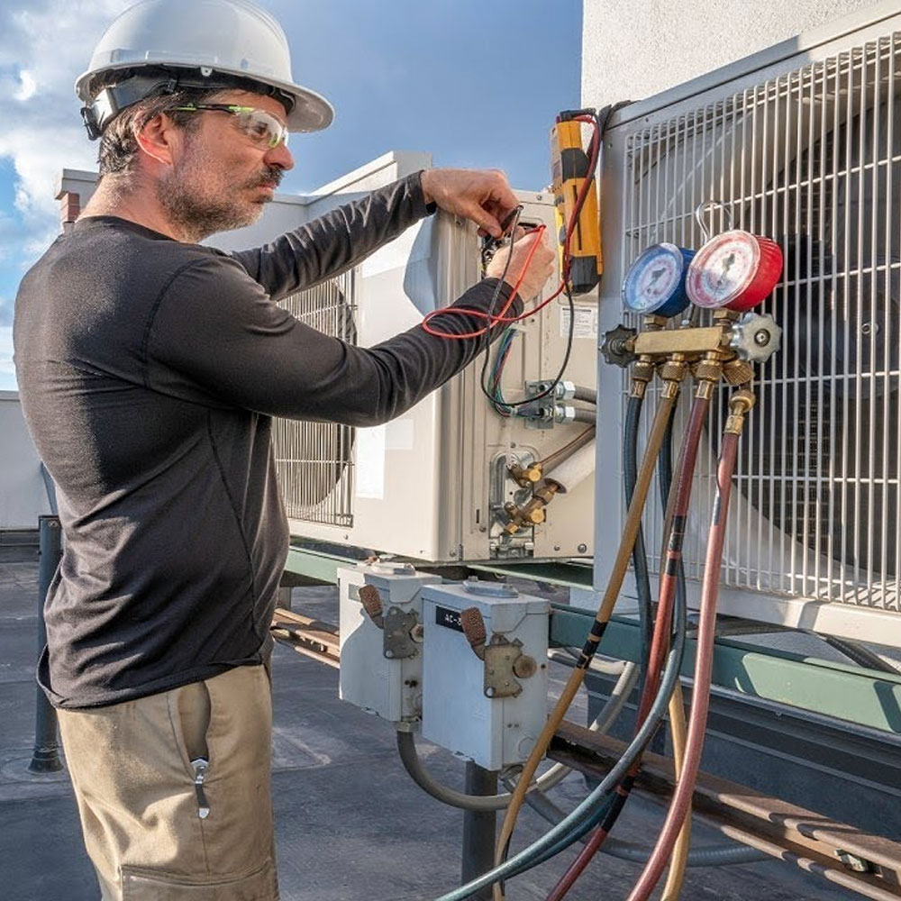 HVAC technician wearing a hard hat and safety glasses using gauges and tools to service an outdoor air conditioning unit