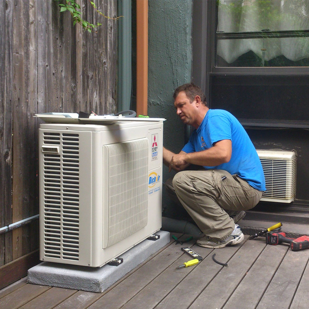 Technician kneeling on a wooden deck and working on an outdoor HVAC unit with tools nearby.