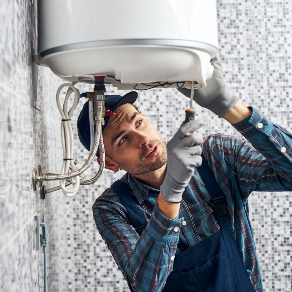 Male plumber in work overalls and gloves repairing a wall-mounted water heater with a screwdriver.